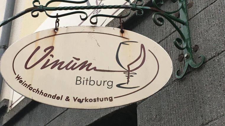 Ein hübsches Schild mit der Aufschrift "Vinum Bitburg". Es zeigt einen Weinfachhandel und eine Verkostung.