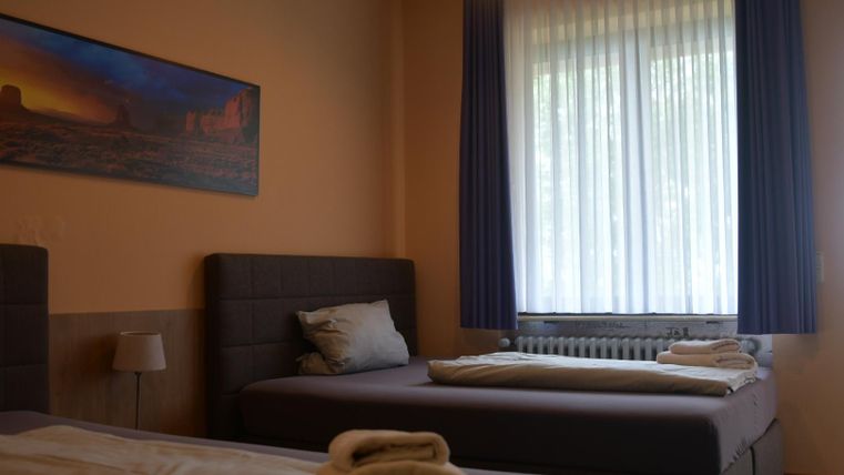 Ein einfaches Zimmer mit zwei Betten und einem Fenster, das viel Tageslicht hereinlässt. An der Wand hängt ein Bild einer Landschaft.