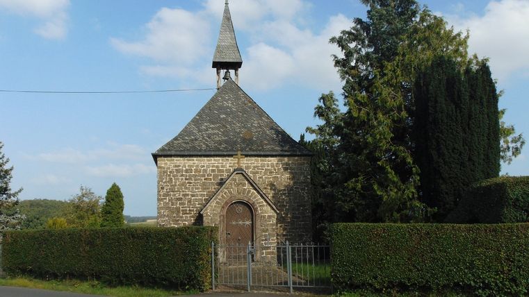 Een kleine stenen kapel met een puntdak en een windhaan, omgeven door bomen en heggen.