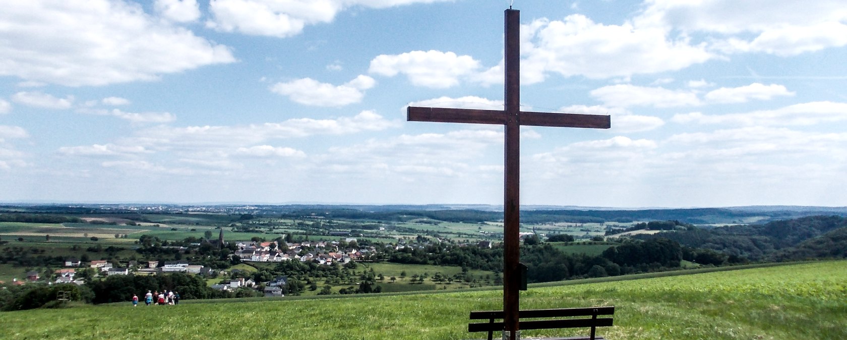 Holzkreuz auf einer Wiese mit Bank, im Hintergrund eine weite Landschaft und ein Dorf unter blauem Himmel mit Wolken., © TI Bitburger Land