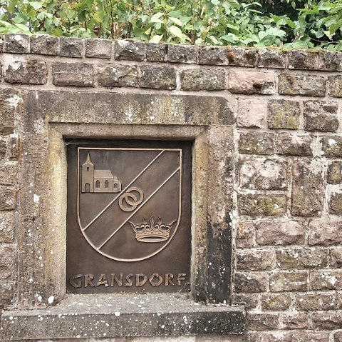 Mur de pierre avec un blason de Gransdorf représentant une église, deux anneaux et une couronne. A droite, on peut voir un tuyau d'évacuation., © TI Bitburger Land S.Wagner