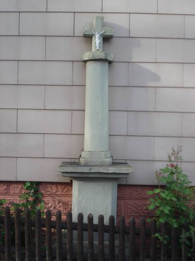 Wegekreuz vor einer Hauswand mit kleinem Holzzaun im Vordergrund.