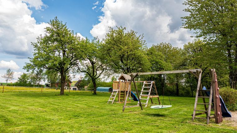 Ein Spielplatz mit einer Schaukel und einem Spielturm in einer grünen Wiese. Im Hintergrund gibt es große Bäume und einen klaren Himmel.
