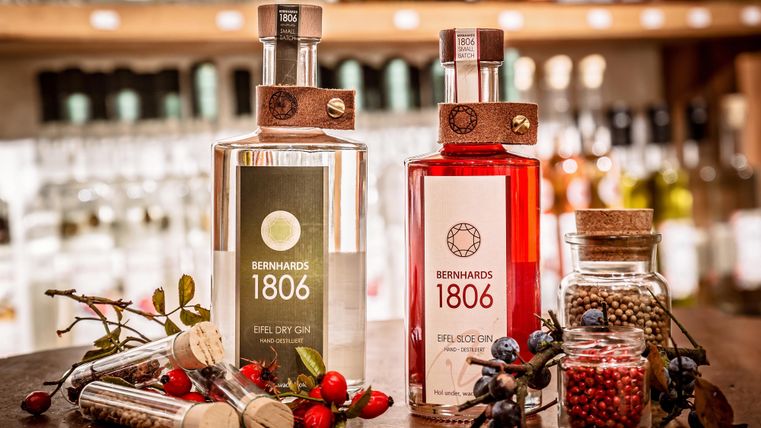 Zwei Flaschen Gin der Marke Bernhards 1806 stehen auf einem Holztisch. Im Hintergrund sind verschwommene Spirituosenregale zu sehen.