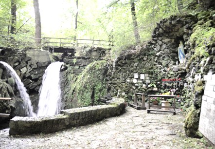 La grotte de Marie d'Albach présente deux chutes d'eau, une statue de la Vierge dans une niche rocheuse et des bougies sur un autel. Entourée d'une végétation luxuriante., © TI Bitburger Land