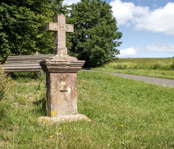 Steinernes Wegekreuz mit 'IHS'-Inschrift auf Wiese, daneben eine Bank, umgeben von Bäumen und einem Weg., © TI Bitburger Land - Monika Mayer