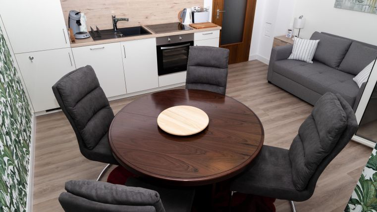 Eine moderne Küche mit einem runden Holztisch und grauen Stühlen. Im Hintergrund befindet sich ein helles Wohnzimmer mit einem Sofa und Pflanzendekoration.