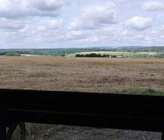 Un banc se trouve devant un vaste champ avec vue sur des collines boisées sous un ciel nuageux., © TI Bitburger Land